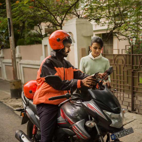man sitting on a motorized bike and holding a mobile phone next to a woman standing and holding a mobile phone